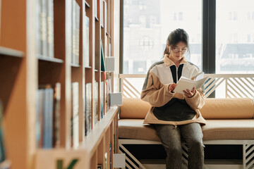 Young serious woman sitting on couch against window and reading new novel of her favorite author in library