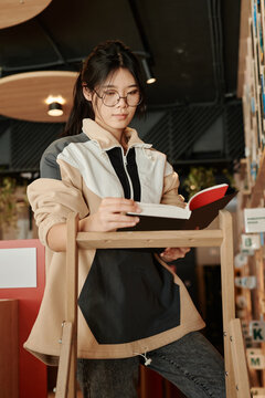 Young erudite in eyeglasses and casualwear standing on stepladder in library or book shop and reading text