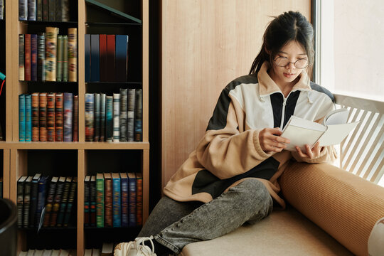 Young Asian student with open book sitting on couch by window in library and preparing for exam before session