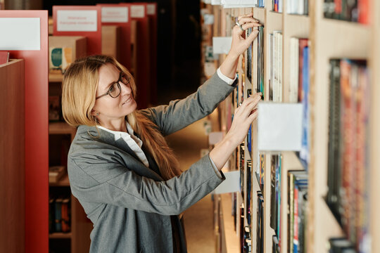 Happy young woman with long blond straight hair standing in front of large stack of shelves in library and looking for book