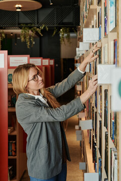 Side view of young blond woman in smart casualwear and eyeglasses seeking for book on shelf in modern library or shop