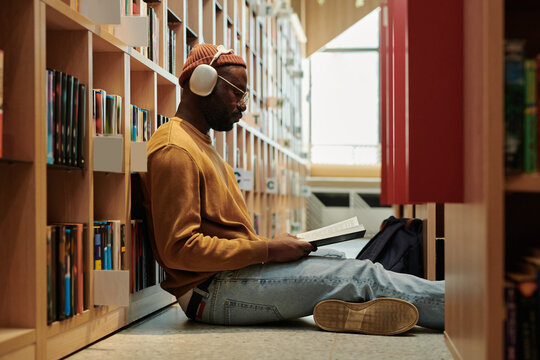 Serious student in headphones sitting on the floor of library in the aisle between stacks of bookshelves and reading