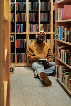 Young man listening to music in headphones while sitting in aisle between bookshelves in library and preparing for exam