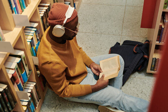 High angle of young African American man in headphones preparing for exam on the floor of library between bookshelves