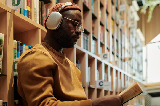 Serious guy in headphones and eyeglasses looking through text in open book while sitting against stacks of bookshelves