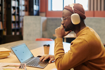 Side view of pensive guy in headphones sitting by table in front of laptop with blank screen in modern library