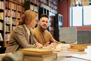 Serious blond woman sitting next to man during discussion of new book at leisure while visiting reading hall of modern library