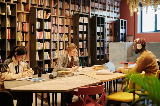 Young intercultural students in casualwear sitting by table in library or reading hall and preparing for exams