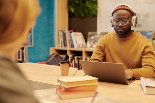 Serious African American guy in eyeglasses, pullover, beanie and headphones networking in front of laptop in library