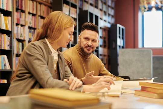 Mature man in quiet luxury attire explaining details of novel plot to blond woman while sitting next to her in reading hall