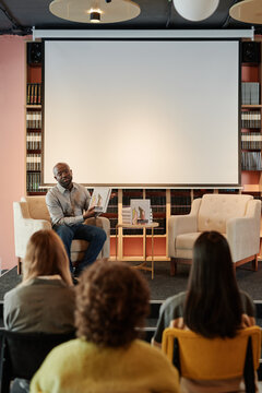 Young confident writer pointing out and describing main points of his new book to public sitting in front of him in library