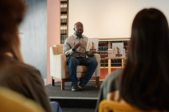 African American author with his new book in hands sitting in armchair in front of public at literature event in modern library