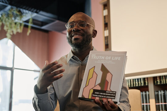 Young African American man in eyeglasses and shirt sitting in front of his fans and presenting new book at literature event