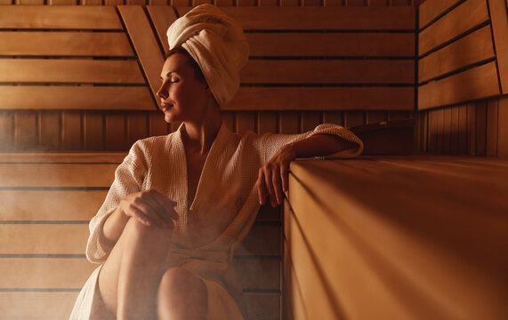 Young woman relaxing and sweating in hot sauna wrapped in towel. Girl In Sauna. Interior of Finnish sauna, classic wooden sauna with hot steam. Russian bathroom. Relax in hot sauna with steam
