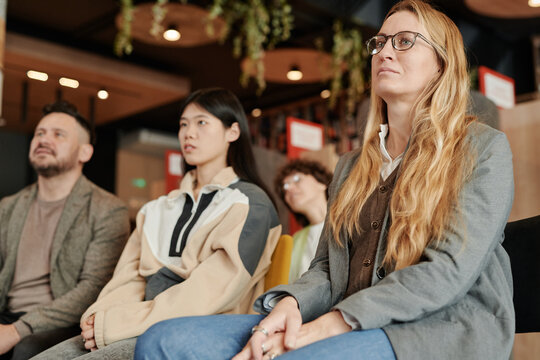 Young serious blond woman in eyeglasses and casualwear sitting next to other people in library and listening to speaker