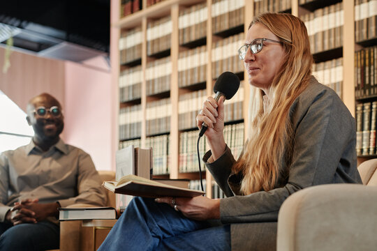 Young female journalist with microphone looking at public during interview with popular modern author in library