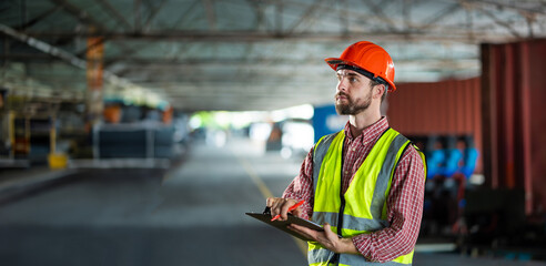 A man in a safety vest is standing in a large empty warehouse. He is holding a clipboard and...
