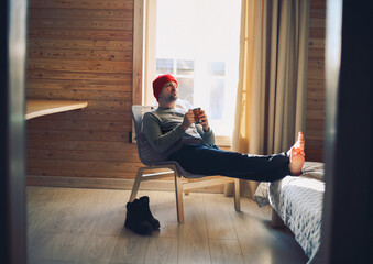 Winter male traveller in red hat holds wooden mug of mulled wine or hot tea with by the window