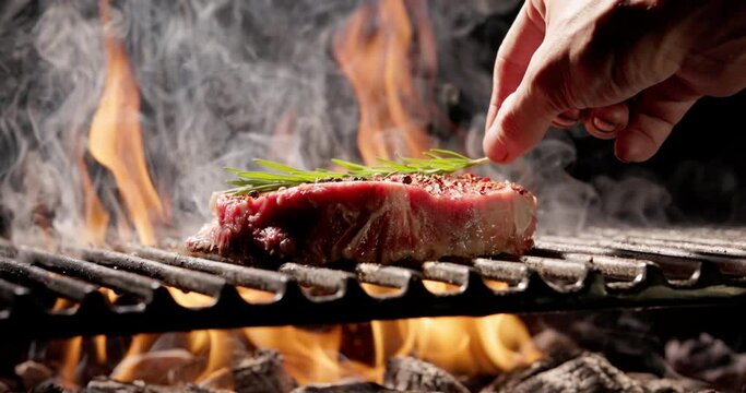 Yellow flames burst out from under grill grate, on which lies juicy piece of steak seasoned with spices, chefs hand gently places sprig of rosemary. Aroma of grilled meat makes your mouth water.