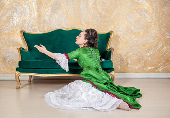 Beautiful sad woman in green rococo style medieval dress sitting on the floor near sofa and raises her hand