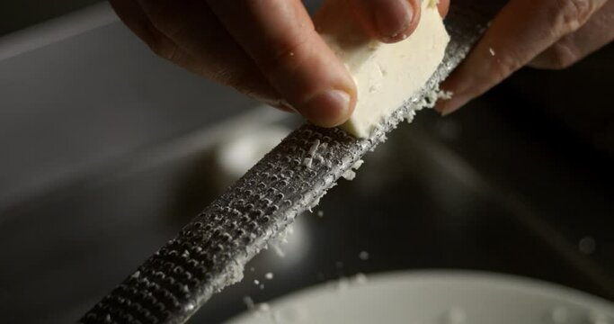 Super slow motion macro of stellar chef grating traditional parmesan cheese with steel grater on the dish close up and pieces of cheese fall at 1000 fps for preparing pasta.
