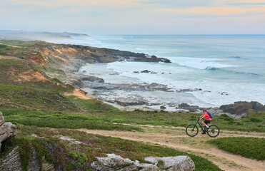 nice senior woman riding her electric mountain bike at the rocky and sandy coastline of the atlantic ocean in Porto Covo, Portugal, Europe
