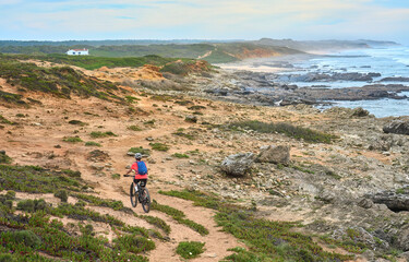 nice senior woman riding her electric mountain bike at the rocky and sandy coastline of the atlantic ocean in Porto Covo, Portugal, Europe - 785612364