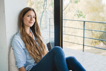 Dreamy young woman sitting by the window thinking about future events at home alone. Beautiful girl female lady dreaming , making plans, creating new ideas