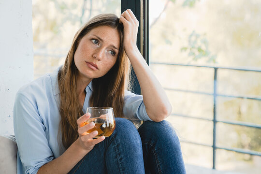 Sad stressed young woman thinking about bad memories drinking juice by the window alone. Confused depressed female girl lady drinking wine and having nostalgic feelings