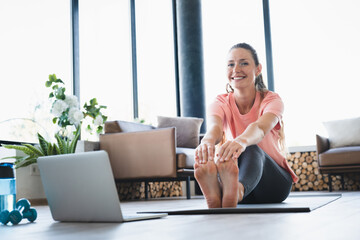 Smiley young Caucasian woman stretching while watching online remote training on laptop at home. Fit female athlete practicing sporty fitness yoga exercises with online tutorials