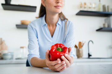 Cropped young Caucasian woman holding red bell pepper standing in the kitchen. Closeup image of...