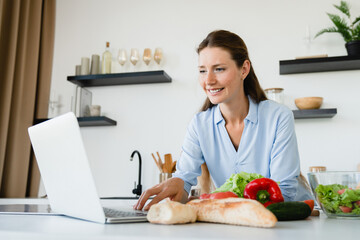 Young Caucasian woman looking for recipe for cooking on laptop at home kitchen. Female housewife chef preparing vegan healthy vegetarian food meal. Healthy eating habits