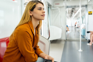 A thoughtful young woman wearing an orange blazer sits inside an empty subway car, looking into the...