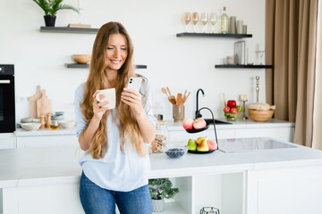 Young woman using cellphone while drinking hot coffee at home kitchen. Caucasian smart girl female freelancer scrolling social media online while having breakfast indoors