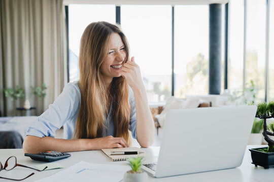Smiley young woman calculating funds and laughing while using laptop at home office. Caucasian freelancer in positive mood paying domestic bills, loans, mortgage, insurance - Powered by Adobe