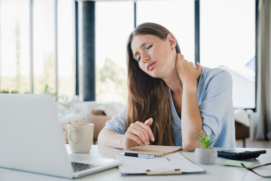 Tired young woman suffering from neck pain while working on laptop at home desk. Freelancer manager office employee rubbing muscles for ache pain relief
