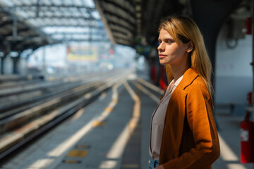 A contemplative young woman stands alone on a sunny train station platform, seemingly waiting for...
