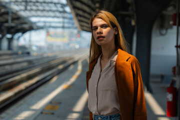 A contemplative young woman stands alone on a sunny train station platform, seemingly waiting for the next train.