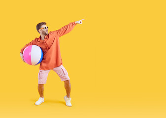 Happy man with inflatable sea ball pointing finger to copy space. Full length portrait of happy young man wearing summer casual clothes and sunglasses having fun on isolated yellow studio background