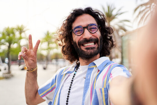 Happy man using phone to take a selfie on vacation, looking at the camera with a toothy smile gesturing the victory sign. Male hippie person doing a photo of himself to post on social media apps.