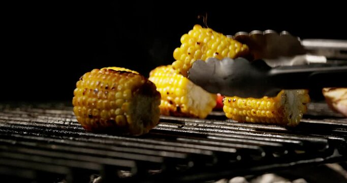 Close-up. Chef turns golden grilled corn with tongs. Grilled vegetables spread delicious aroma through air. Golden corn, aromatic garlic and cherry tomatoes are excellent addition to meat. Copy space