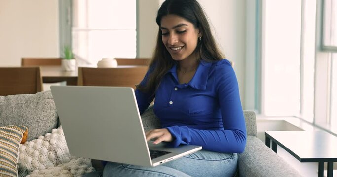 Happy young Indian woman using laptop on comfortable home couch, working on freelance project, sitting on sofa, typing, turning look away, thinking on creative ideas, decision for Internet startup