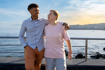 Two friends, male and non-binary, share laughter on a seaside promenade at sunset, with the ocean...