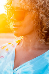 Sunny outdoor portrait of curly young woman smiling and wearing sunglasses with sunset backlight. Golden portrait of attractive lady in outdoors leisure activity alone. Tourist happy. Beautiful female