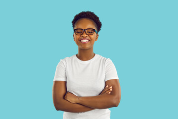 Positive young African American woman smiling broadly with arms crossed in front of chest confidently looking at camera wearing glasses and white casual T-shirt stands on turquoise studio background