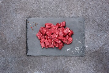 Sliced raw beef meat for slices for the preparation of goulash, stew on a shaleboard on a black concrete background.