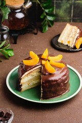 A biscuit cake with a cream of masquearpone, canned peaches covered with chocolate on a green plate on a brown concrete background.
