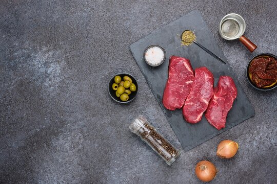 Prepared ingredients for cooking stewed beef with dried tomatoes and olives on a black concrete background.