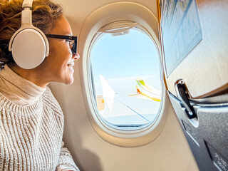 Smiling passenger with Bluetooth noise-canceling headphones looking out of the airplane's porthole...