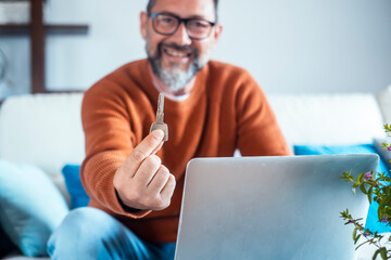 Man with glasses sitting on the sofa at home in front of his laptop smiling showing the key to his...